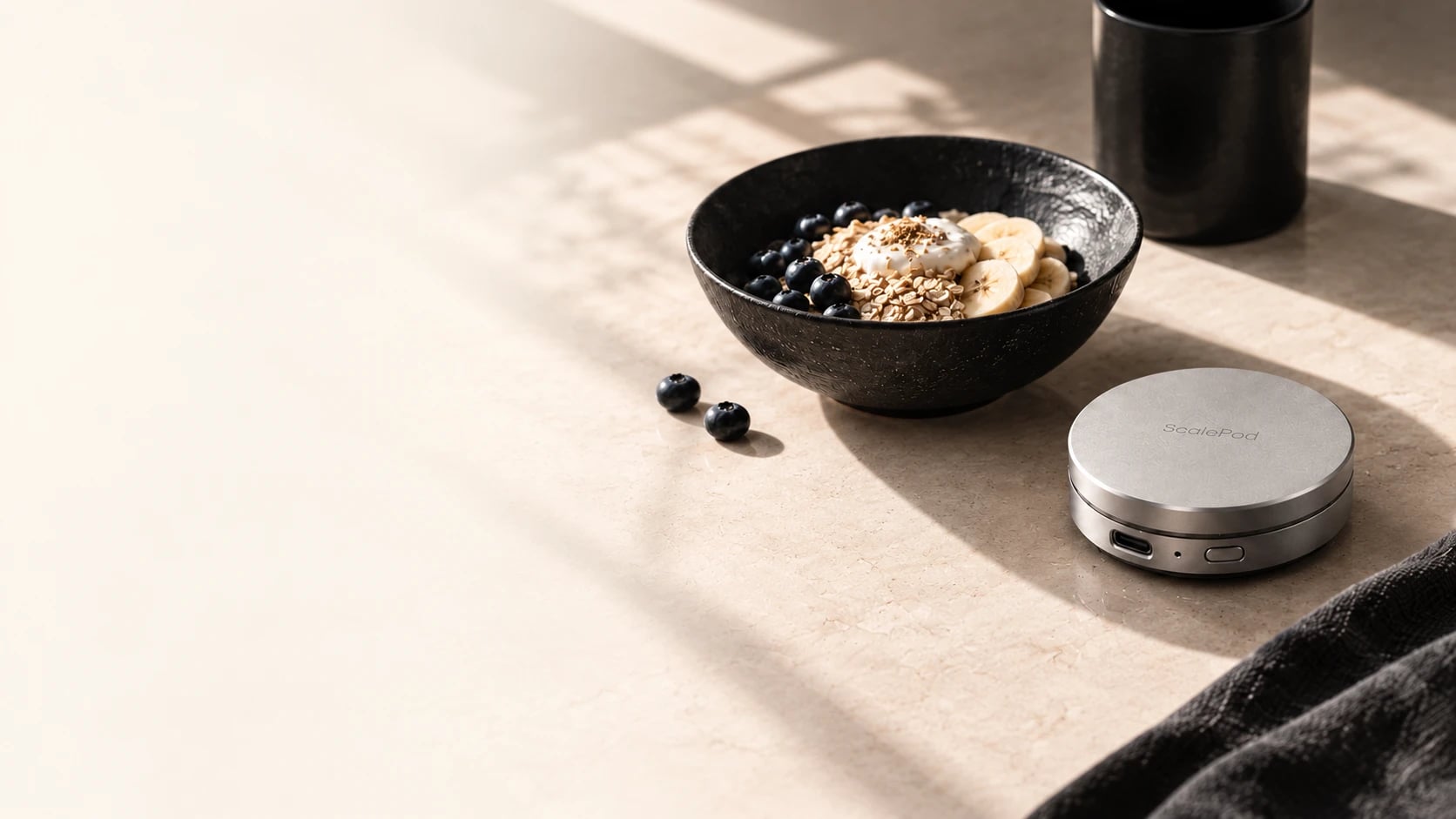 A black stone bowl of oats, yogurt, blueberries and banana beside a ScalePod pocket scale on a sunlit cream stone counter.